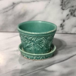 May include: A teal ceramic planter with a matching saucer. The planter features a detailed embossed design of leaves and dots around the exterior. The small, round saucer has a similar embossed pattern. The planter is designed for small plants.