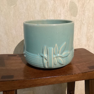 May include: A cylindrical ceramic planter in a light turquoise blue glaze. The exterior features a raised relief design of bamboo stalks and leaves encircling the lower portion, complemented by horizontal grooved lines around the base.