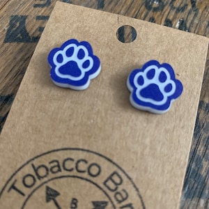 May include: A pair of blue and white paw print stud earrings. The earrings are on a brown cardboard backing with the words "Tobacco Barn" printed on it. The earrings are small and have a white paw print design on a blue background.