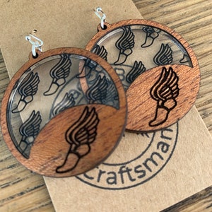 May include: A pair of wooden earrings with a clear acrylic insert featuring a winged foot design. The earrings are brown and have silver hooks. The earrings are displayed on a brown paper card with the word "craftsman" printed on it.