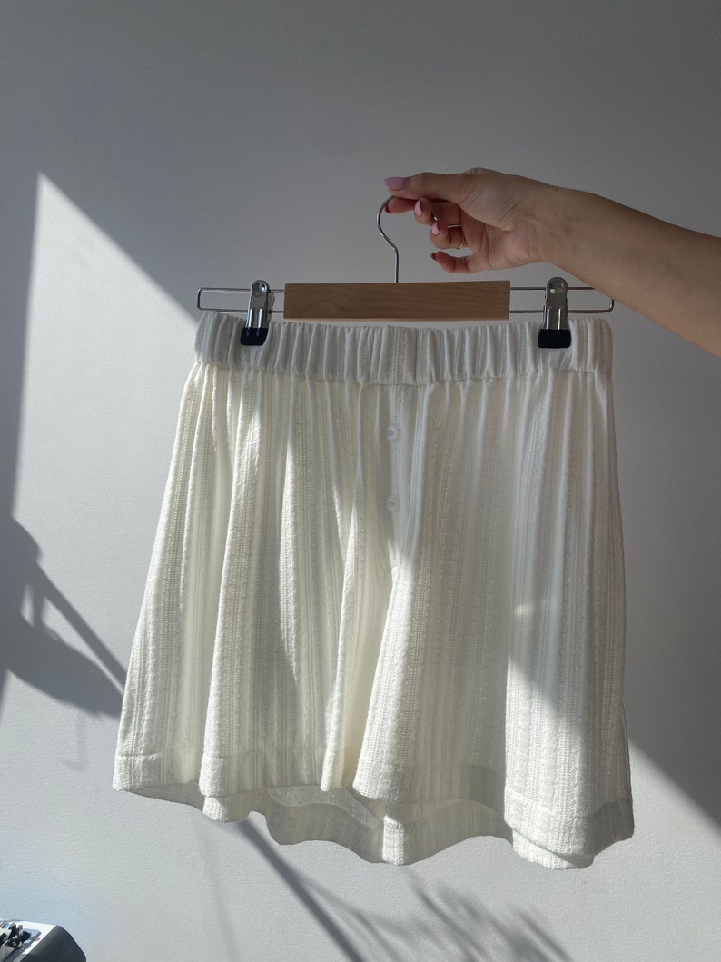 May include: White cotton shorts with a button fastening and a relaxed fit. The shorts are hanging on a wooden hanger.
