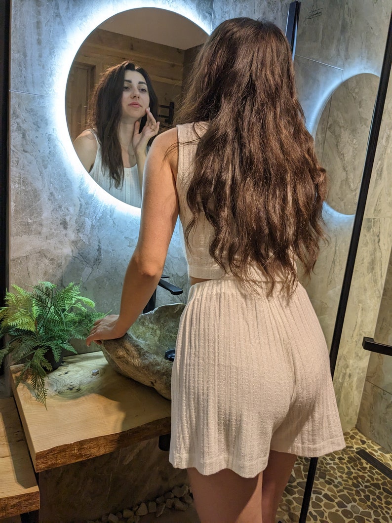 May include: A woman wearing a white ribbed tank top and matching shorts stands in front of a bathroom mirror. She is looking at her reflection in the mirror.