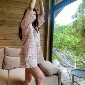 Peut inclure: Une femme portant un pyjama rose et blanc &agrave; motifs avec des manches courtes et un short. Elle se tient dans une pi&egrave;ce avec un mur en bois et une grande fen&ecirc;tre donnant sur une for&ecirc;t verte.