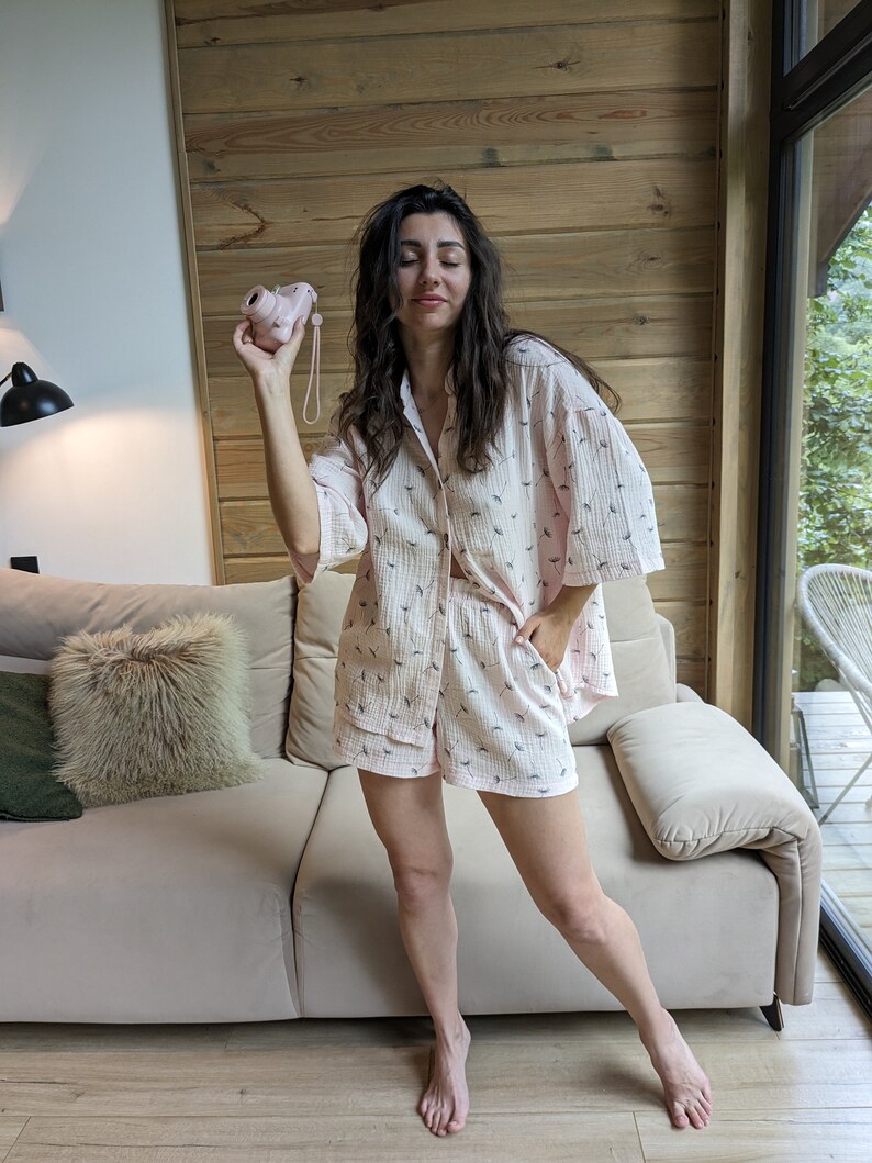 Peut inclure: Une femme portant un pyjama rose et blanc &agrave; motifs avec des manches courtes et un short. Elle tient un appareil photo jouet rose dans sa main droite et se tient devant un canap&eacute;.