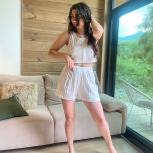 May include: A woman wearing a white striped crop top and matching shorts. She is standing in a room with a wooden wall and a large window.