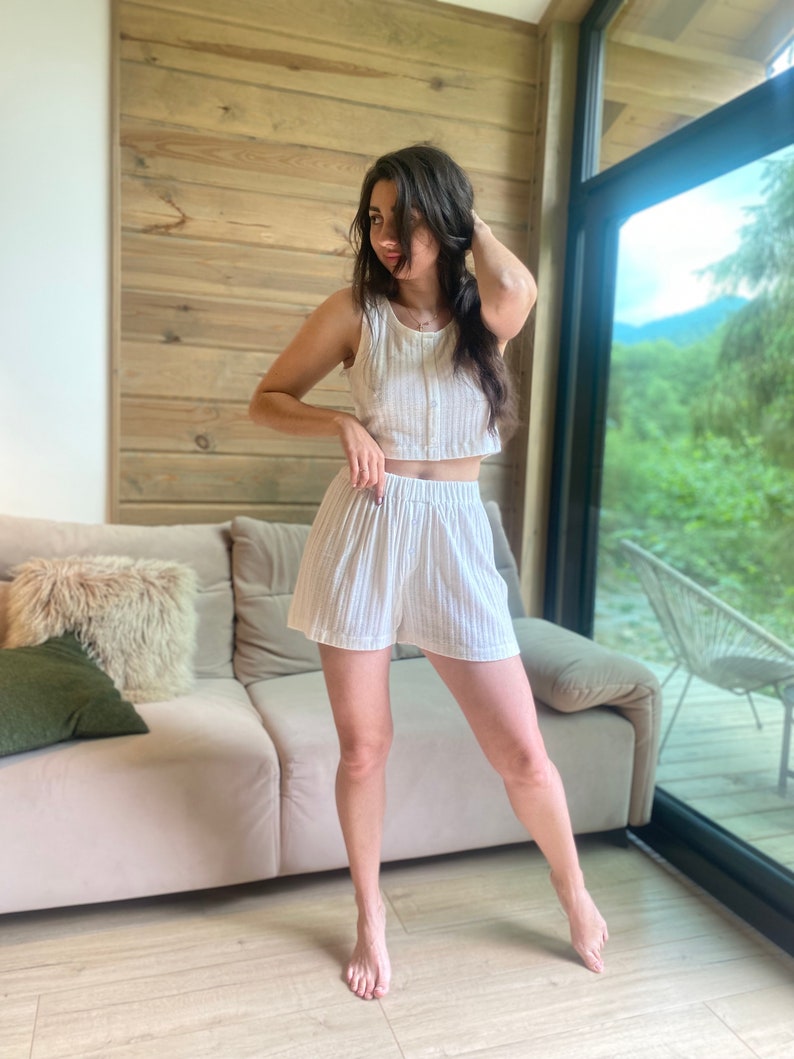 May include: A woman wearing a white striped crop top and matching shorts. She is standing in a room with a wooden wall and a large window.