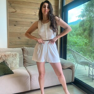 May include: A white sleeveless crop top and matching shorts set with a striped pattern. The model is standing in a room with a wooden wall and a window.