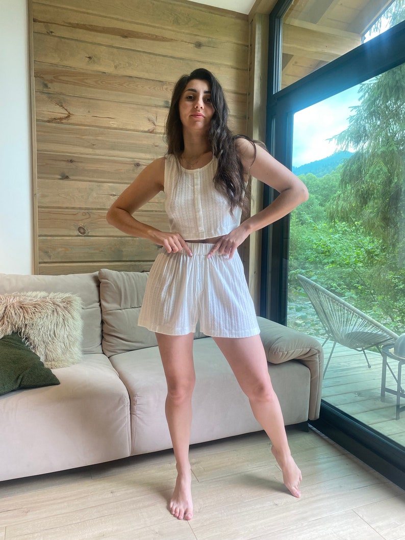 May include: A white sleeveless crop top and matching shorts set with a striped pattern. The model is standing in a room with a wooden wall and a window.