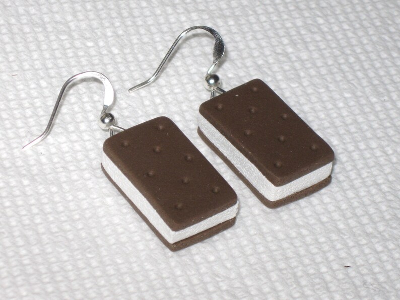May include: A pair of silver dangle earrings with a brown and white ice cream sandwich charm. The charm is made of polymer clay and has a realistic look.