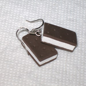 May include: A pair of earrings shaped like ice cream sandwiches. The earrings are made of brown and white polymer clay and have silver hooks.