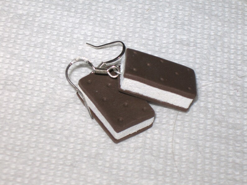 May include: A pair of earrings shaped like ice cream sandwiches. The earrings are made of brown and white polymer clay and have silver hooks.