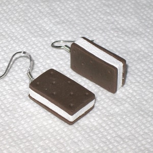 May include: A pair of dangle earrings featuring a chocolate and white ice cream sandwich design. The earrings are made of polymer clay and have silver hooks.