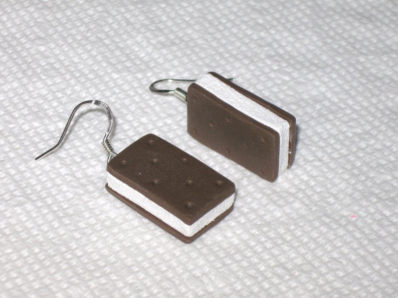 May include: A pair of dangle earrings featuring a chocolate and white ice cream sandwich design. The earrings are made of polymer clay and have silver hooks.