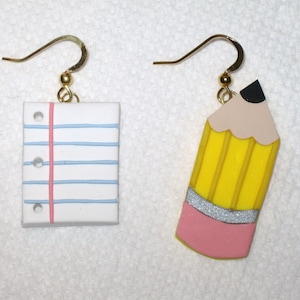 May include: A pair of earrings featuring a yellow pencil with a pink eraser and a white sheet of paper with blue lines and a pink line.