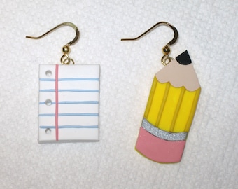 May include: A pair of earrings featuring a yellow pencil with a pink eraser and a white sheet of paper with blue lines and a pink line.
