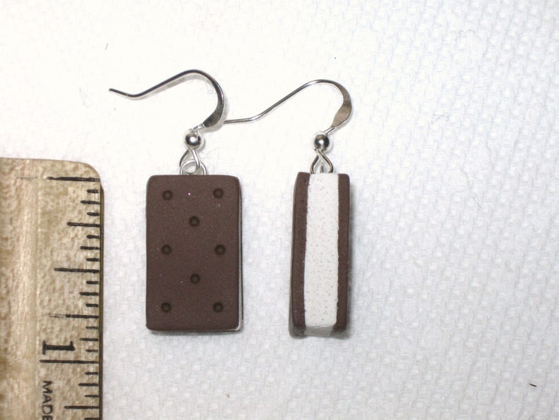May include: A pair of earrings featuring a chocolate and vanilla ice cream sandwich design. The earrings are made of polymer clay and have silver hooks.