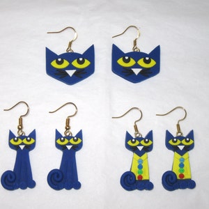 May include: A set of six blue cat earrings. Two earrings feature a simple cat face design, two feature a cat with a curled tail, and two feature a cat wearing a yellow shirt with colorful buttons.