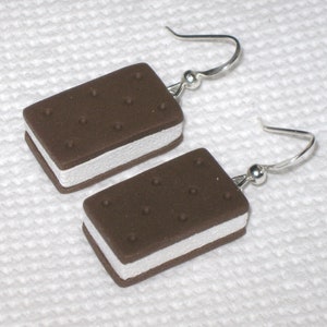 May include: A pair of earrings shaped like ice cream sandwiches. The earrings are made of polymer clay and have a brown chocolate cookie exterior with a white vanilla ice cream filling. The earrings are attached to silver hooks.