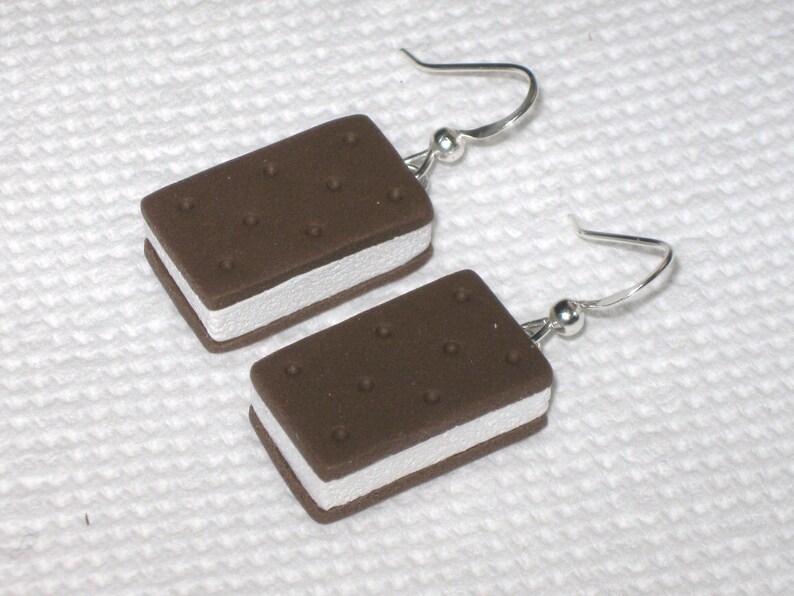 May include: A pair of earrings shaped like ice cream sandwiches. The earrings are made of polymer clay and have a brown chocolate cookie exterior with a white vanilla ice cream filling. The earrings are attached to silver hooks.