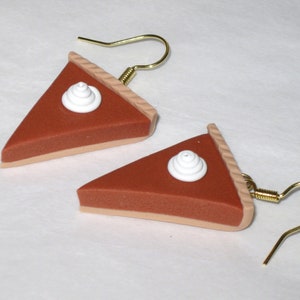 May include: A pair of dangle earrings shaped like pumpkin pie slices with white whipped cream on top. The earrings are made of polymer clay and have gold hooks.