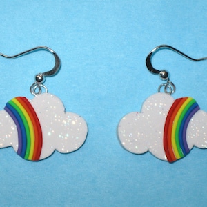 May include: A pair of silver dangle earrings featuring a white cloud with a rainbow design. The cloud is covered in glitter.