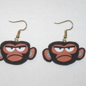 May include: A pair of earrings featuring cartoon monkey faces with angry expressions. The monkeys have black fur, brown skin, and white eyes. The earrings are made of gold-colored metal.