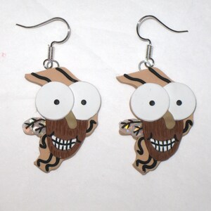May include: A pair of dangle earrings featuring a cartoon character with large eyes, a brown beard, and a toothy grin. The character is depicted in a tan and brown color scheme with black accents.