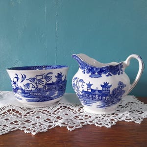 Puede incluir: Un juego de azucarero y jarra de crema de porcelana azul y blanca con un diseño chino tradicional. El juego presenta un delicado patrón floral y una escena de un puente y una pagoda.