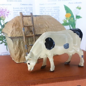 May include: A white cow with black spots is standing next to a small thatched roof hut with a ladder. The cow is looking down at the ground. The hut and cow are made of painted metal.
