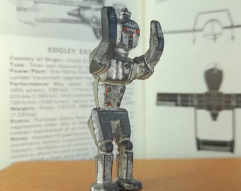 Könnte beinhalten: Kleine, silberfarbene Roboterfigur mit erhobenen Armen. Der Roboter hat rote Augen und steht vor einem offenen Buch mit Text und Illustrationen. Das Design des Roboters ist einfach, mit einem kantigen Körper und Beinen.
