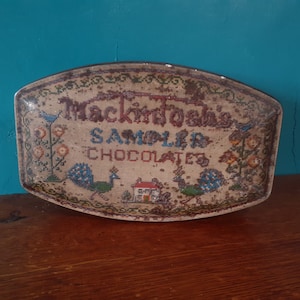 Könnte beinhalten: Eine antike Blechdose mit einem Kreuzstich-Design mit dem Text "Mackintosh's Sampler Chocolates". Das Design enthält auch zwei Pfauen und ein kleines Haus.
