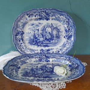 May include: Two oval ceramic platters featuring a blue and white transferware pattern. The design depicts classical architectural ruins, trees, and water scenes. The scalloped edges are decorated with floral motifs. A single white rose rests on one.