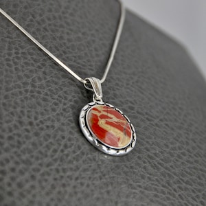 May include: Oval pendant necklace with a silver chain. The pendant features a red and beige stone set in a silver bezel with a decorative edge. The necklace is displayed on a gray surface.