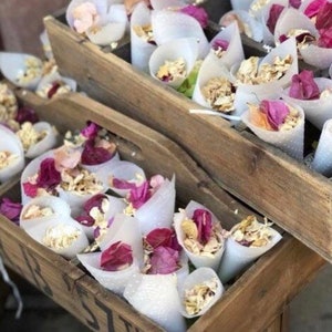May include: Two wooden crates filled with white paper cones filled with dried flower petals. The cones are tied with twine and the petals are pink and white.
