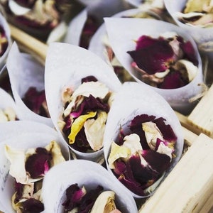 May include: White paper cones filled with dried rose petals in shades of pink, red, and white. The cones are arranged in a wooden box.