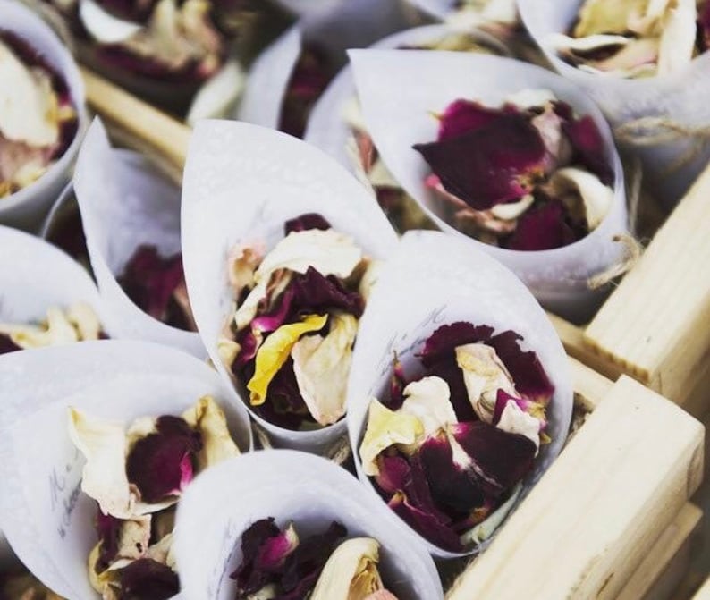 May include: White paper cones filled with dried rose petals in shades of pink, red, and white. The cones are arranged in a wooden box.