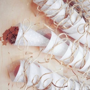 May include: White paper cones with brown twine tied around them, filled with brown confetti stars. The cones are arranged on a light brown wooden surface.