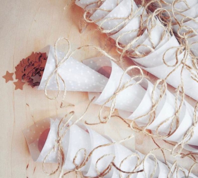 May include: White paper cones with brown twine tied around them, filled with brown confetti stars. The cones are arranged on a light brown wooden surface.