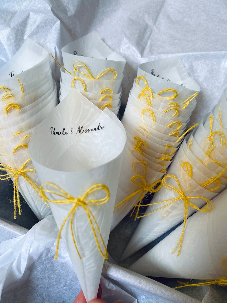 May include: A stack of white paper cones with yellow string bows. The cones are decorated with a delicate floral pattern and the text "Pamela & Alessandro".