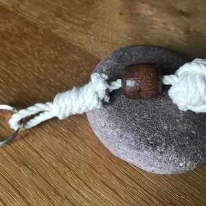 May include: A white rope keychain with a brown wooden bead and a knot at the end. The keychain is attached to a silver key ring.