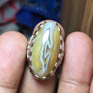 May include: An oval-shaped ring featuring a polished, translucent yellow and white banded agate gemstone set within a silver-toned metal prong setting. The stone displays natural swirling patterns and light reflections across its smooth surface.