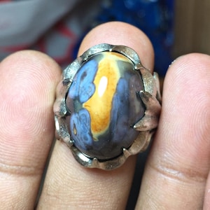 May include: A silver-toned ring featuring a large, oval-shaped cabochon stone with a smooth, polished surface. The stone displays a central vertical band of golden yellow surrounded by swirling patterns of deep blue, grey, and translucent brown.
