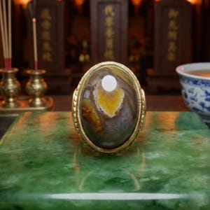 Op de afbeelding: Een gouden ring met een grote, ovaalvormige cabochonsteen met een glad, gepolijst oppervlak. De steen vertoont een gemarmerd patroon van diepbruin, grijs en een centrale vlek van levendig geel. De metalen band heeft een getextureerde rand.