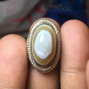 May include: An oval ring featuring a polished, banded stone with a white center surrounded by a tan ring, set in a silver-toned metal band with a textured, notched edge detail around the bezel. The stone has a smooth, glossy, reflective surface.