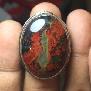 May include: An oval-shaped ring featuring a polished gemstone with a mottled pattern of deep red, dark green, and black inclusions, set within a simple silver-toned metal band that encircles the stone's perimeter with a smooth, reflective finish.