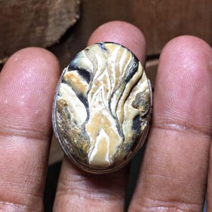May include: An oval-shaped ring featuring a polished stone with a swirling pattern of cream, tan, brown, and black colors. The stone is set within a simple silver-toned metal band that encircles the perimeter of the gemstone.