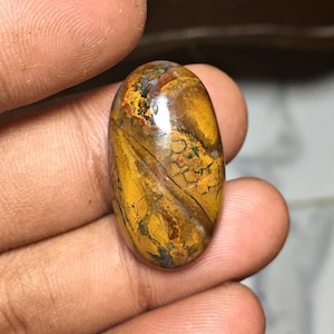 May include: An oval-shaped polished gemstone featuring a complex pattern of mustard yellow, deep brown, and dark grey veins. The surface is smooth and reflective, displaying a natural matrix with intricate, interconnected lines and earthy tones.