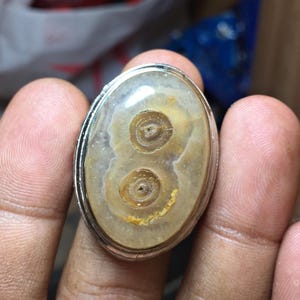 Può includere: Un anello di forma ovale con una pietra lucida e traslucida caratterizzata da due distinte inclusioni circolari color marrone, simili a occhi, incastonata in una fascia di metallo color argento liscio con un motivo marmorizzato beige.