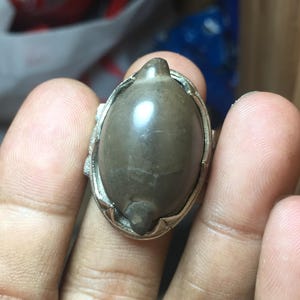 May include: A ring featuring a smooth, oval-shaped, dark brown stone set within a silver-toned metal frame. The stone has a polished, reflective surface with a subtle, natural texture. The metal setting has a decorative, notched edge design.