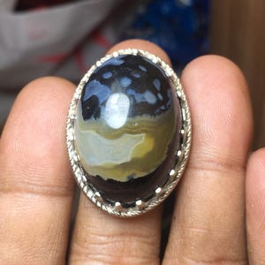 May include: An oval-shaped cabochon gemstone ring set in a silver-toned metal band with a twisted rope-style border. The stone features a dark black base with swirling patterns of translucent gray, white, and muted yellow tones throughout.